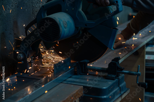 Worker cutting metal profile with electric miter saw, flying sparks in industrial metal fabrication workshop.