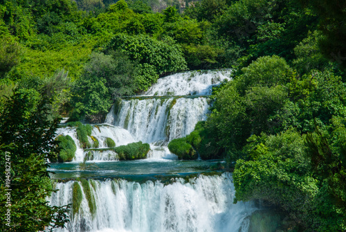 Breathtaking multi-tiered waterfall surrounded by dense green foliage, with cascading water flowing over vibrant, moss-covered rocks into lake. Krka National Park is of Croatia's natural treasures.