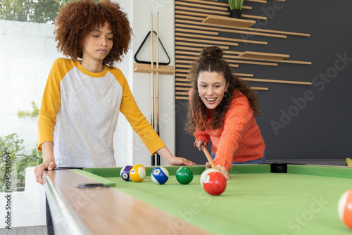 Diverse female friends shooting with cue stick at pool table in game room, wearing orange sweater