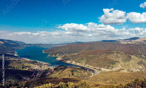 View of the tranquil lake reflecting the azure sky, embraced by rolling hills of lush forest under a blanket of fluffy clouds, Mavrove, North Macedonia.
