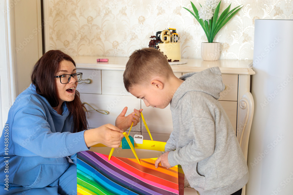 Obraz premium excited mother watching little boy opening colorful birthday gift bag during sixth birthday celebration at home with construction themed cake on background