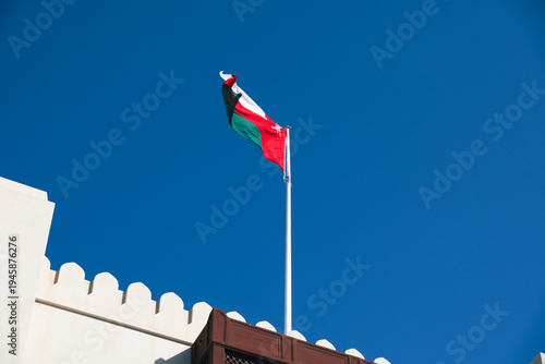 Fotografia Oman national flag waving against clear blue sky above traditional Governate bui