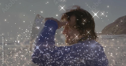 Vertical video: Sun glaring causing surfer shielding eyes, holding board on beach, wetsuit, sparkle