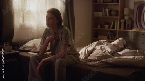 Young adult woman sitting on bedside pressing hand to chest struggling with panic attack feeling shortness of breath then drinking water to calm down in dimly lit bedroom