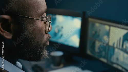Side view close up shot of African American man working as security officer and answering phone calls while reviewing CCTV footage on multiple computer monitors during night shift in office