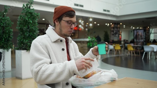 Young frustrated man drinking coffee in a shopping mall food court, checking his shopping receipt and becoming shocked by the high prices, illustrating financial stress and inflation