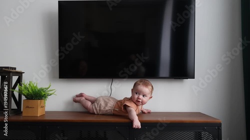 Small child lies on a dark wooden cabinet in a bright room, looking into the camera lens and smiling slightly
