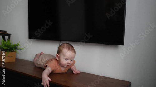 A baby in a brown bodysuit lies on a wooden chest of drawers under a TV, looking around and moving his hands