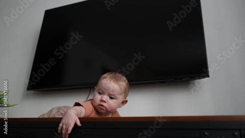 Baby lying on a dark wooden cabinet under a TV, reaching out with one hand and looking down.