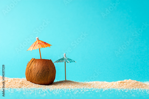 Minimalist summer concept with two coconuts and a cocktail umbrella on white sand and blue background