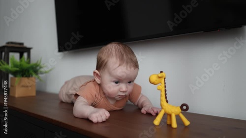 Infant lying on a wooden surface looks at a yellow giraffe toy and starts drooling