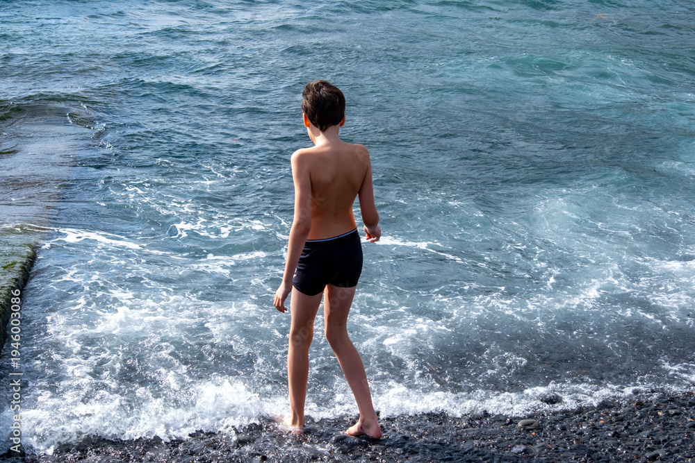 Fototapeta premium Teen boy playing in ocean waves on black volcanic sand beach, joyful summer moment, natural fun, healthy childhood, barefoot freedom