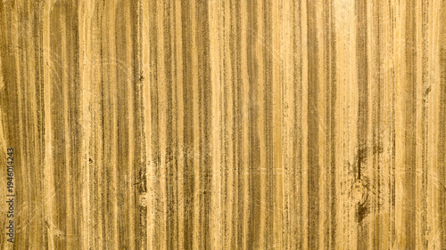 Texture of a harvested golden field seen from directly above. The vertical lines created by farming machinery form a rhythmic, organic pattern across the dry earth, showcasing agricultural geometry.