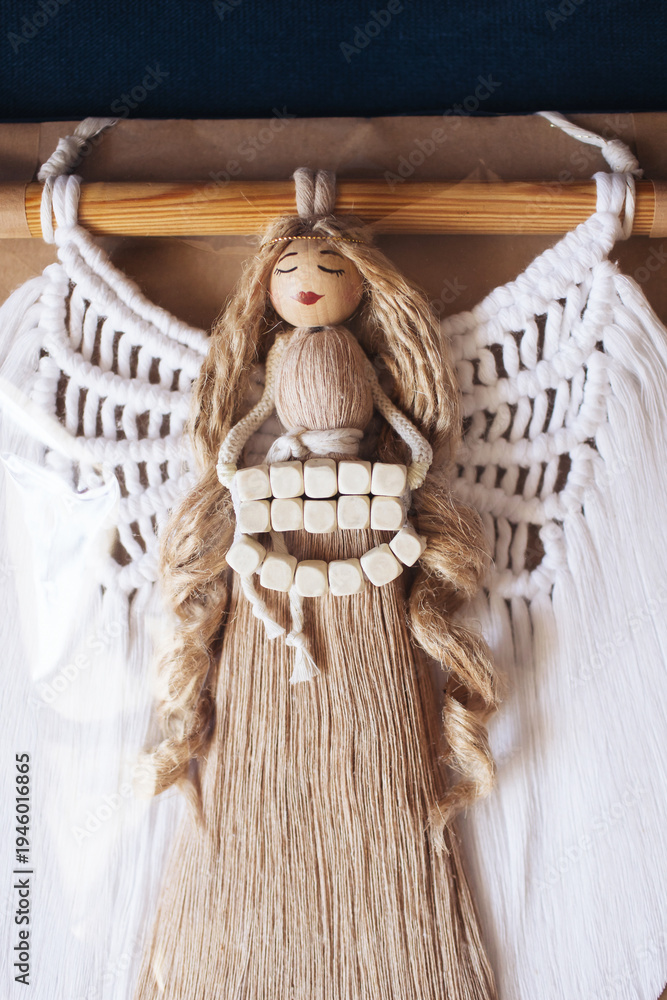 Obraz premium Handmade macrame angel wall decoration with wooden beads and natural cotton cord. Boho craft guardian angel ornament hanging on wooden rod. Rustic handmade spiritual home decor symbol.