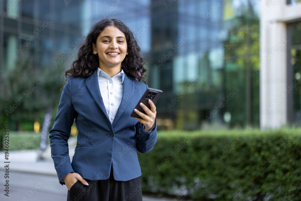 Fototapeta premium Young businesswoman with dark curly hair smiles confidently, holding a smartphone with one hand in pocket before a modern glass office building, symbolizing ambition and digital connectivity