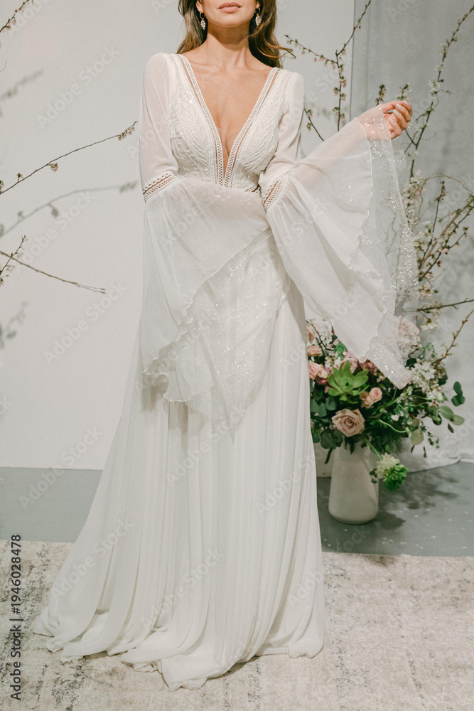 Fototapeta premium Woman in white dress stands in front of floral arrangement, showcasing bridal fashion in a soft light environment during a wedding event