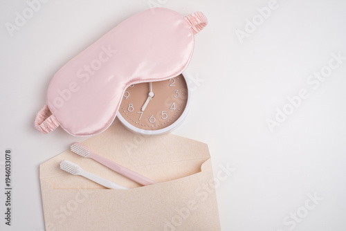 Pink sleep mask, alarm clock, and toothbrushes in envelope on white background. Morning and evening routine concept. 