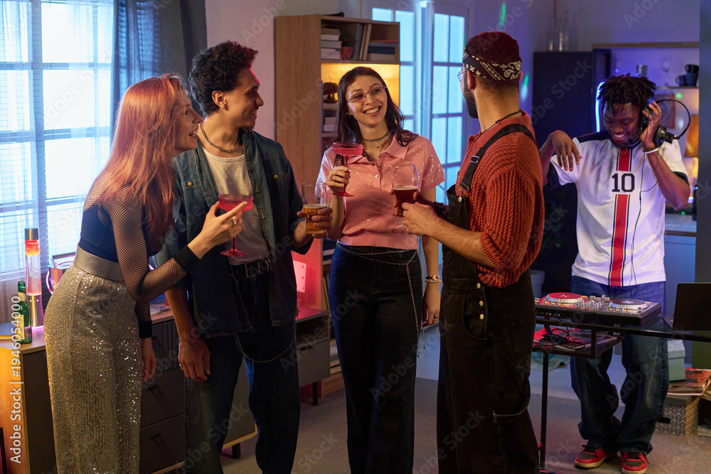 Fototapeta premium Group of multiethnic young people socializing and holding drinks at party while Black man DJing in background, everyone smiling and engaging in lively conversation indoors