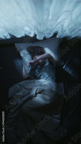 Vertical shot of young adult woman nervously rolling over in bed trying to get comfortable while struggling to fall asleep during night in bedroom
