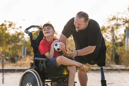 Foto Adult male with a prosthetic limb engaging with a young boy using a wheelchair and holding a soccer ball
