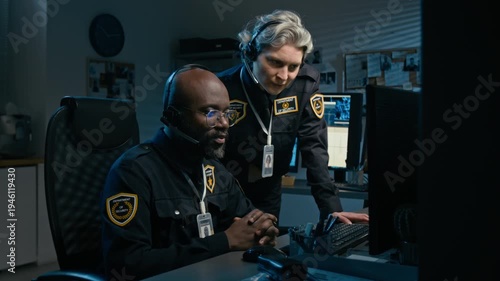 Medium shot of two police officers working together at station during night shift and using computer while pointing at monitor and coordinating operations, copy space