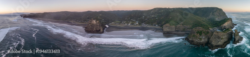 Photography A panoramic view of Piha Beach, New Zealand, showcasing the rugged coastline, lush hills, and crashing waves at sunset