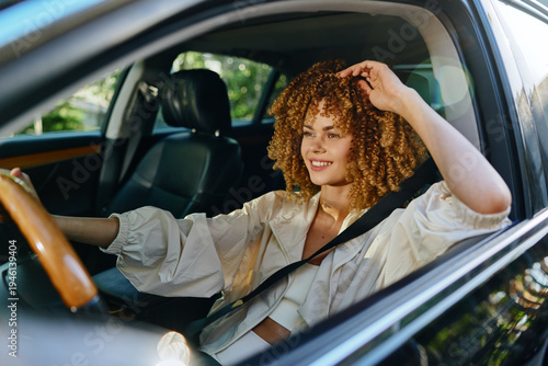 Obraz na płótnie Smiling woman driving a car with curly hair and casual white shirt on a sunny day, enjoying a relaxed moment while seated in a modern vehicle interior