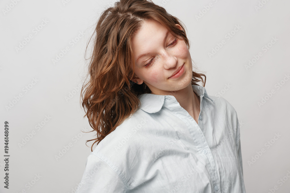 Obraz premium Young woman with wavy hair wearing white shirt looks down with gentle smile on light background. Concept of natural beauty, calmness, and soft emotions in portrait photography.