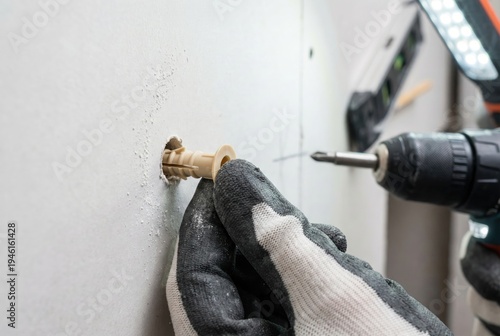 Hand pressing a beige plastic wall anchor into a drilled hole in white drywall with a cordless drill nearby
