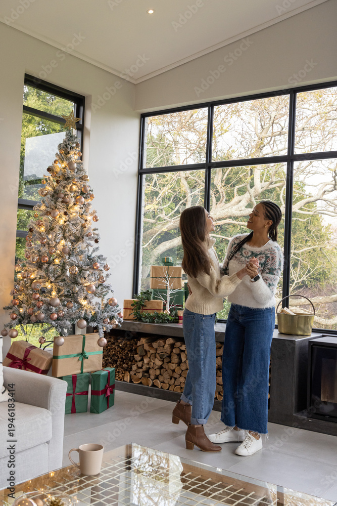 Fototapeta premium Female partners holding hands and swaying in living room by window with decorated Christmas tree