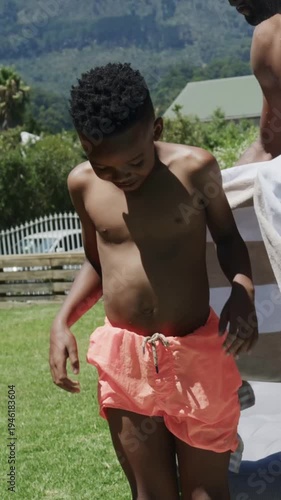 Vertical video: Coming right, dad draping towel over boy in coral shorts on lawn to dry after swim