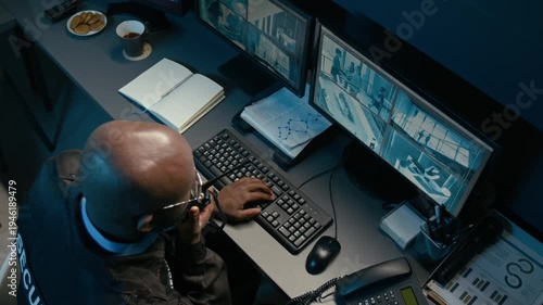 Top view shot of Black man watching computer monitors with CCTV footage while using portable radio and coordinating security operations from control room workstation during night shift