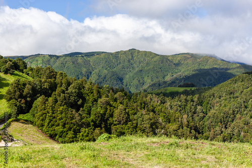 Lush green landscape near Furnas, Sao Miguel. Rolling hills and dense forest under a partly cloudy sky. Volcanic terrain with vibrant greenery and distant mountains.