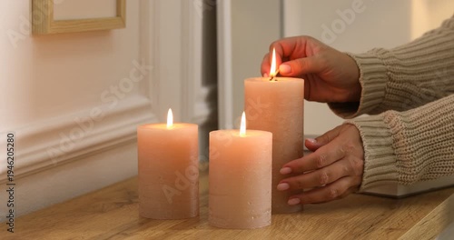 Woman lightning up candle at wooden chest of drawers with books near white wall indoors, closeup