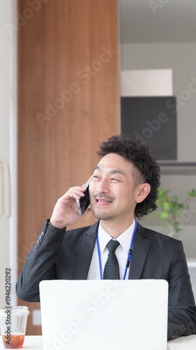 ノートパソコンを使いスマホで電話をするビジネスマンの髭の男性の縦動画