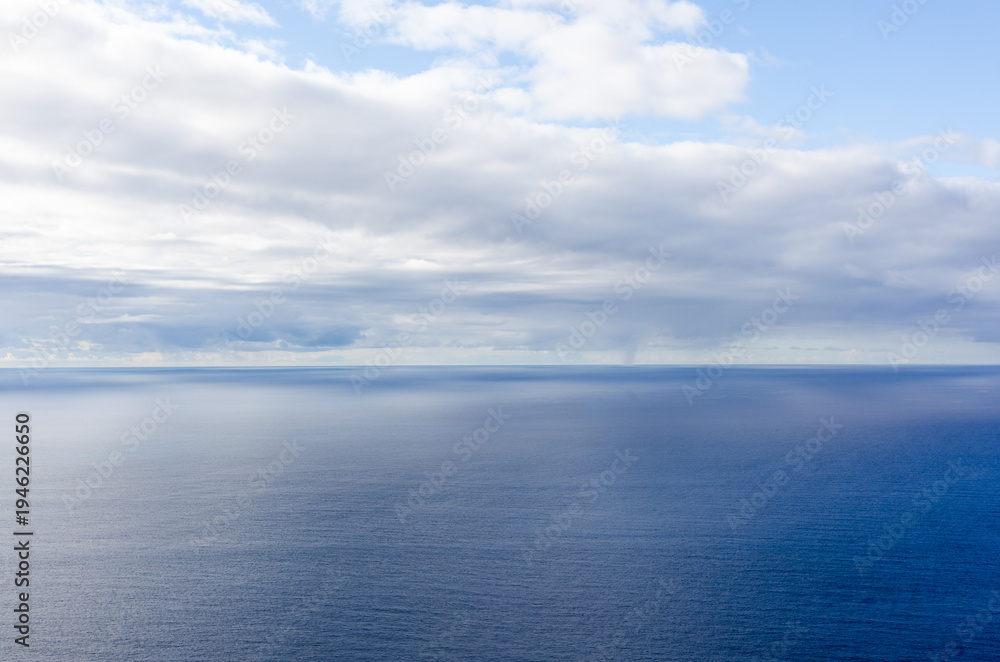 Fototapeta premium Atlantic Ocean view. Calm blue water stretches to the horizon. Low clouds hover above the sea. Scene photographed near Madeira, Portugal.