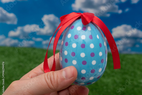 Hand holding a decorated blue Easter egg with a red ribbon outdoors.