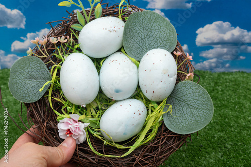 Hand holding a bird's nest with light blue speckled eggs outdoors.