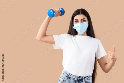 Young woman in medical mask with dumbbell showing thumb-up on beige backgroun...