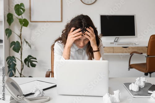 Stressed young businesswoman with laptop and crumpled paper at her workspace ...