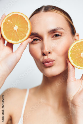 Young woman holding two orange halves near face, making a playful kiss expression, fresh citrus fruit, natural skincare concept, bright background, close-up portrait