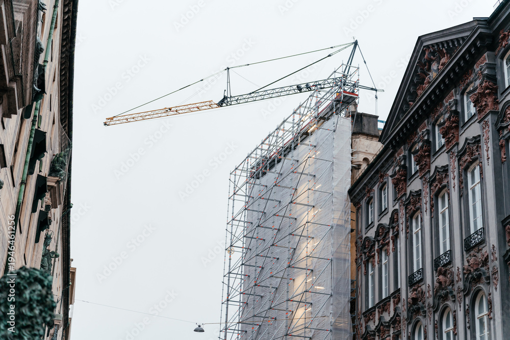 Fototapeta premium Urban construction scene featuring a historic building covered in scaffolding with a crane above, development and architectural transformation.