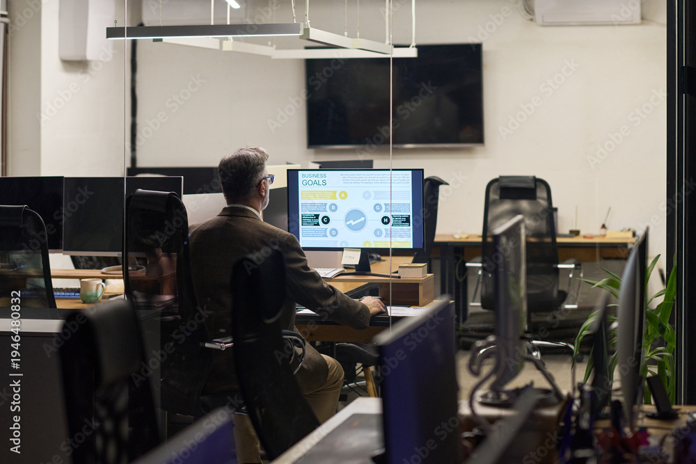 Fototapeta premium Middle aged Caucasian man sitting at desk working on computer with business analytics displayed on monitor in modern office environment, empty workstations in background