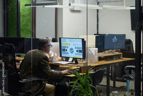 Middle aged Caucasian man working at desk using desktop computer with business analytics displayed on screen in modern office environment, sitting alone in workspace