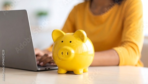Yellow piggy bank sits on desk next to a laptop computer and blurred person typing representing saving money and online finance management