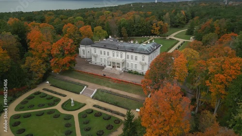 Autumn foliage surrounds a grand white manor and manicured gardens, overlooking a tranquil bay.