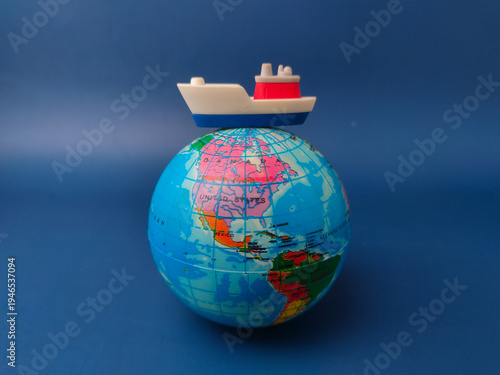 A conceptual still life featuring a miniature toy cargo ship positioned on top a globe on a blue background