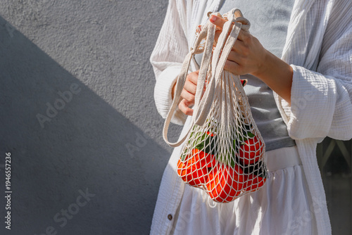 Woman adjusting mesh bag of oranges near textured gray wall in sunlight