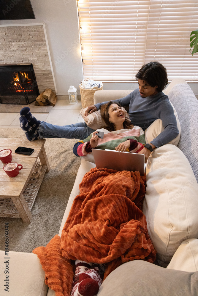 Obraz premium Diverse couple relaxing on light couch in living room, woman holding laptop, fireplace crackling