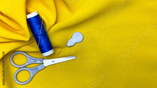 Scissors, a spool of blue thread, and a needle on yellow fabric. Concept art for handicrafts, sewing, clothing repair, home crafts, and sewing tools.
